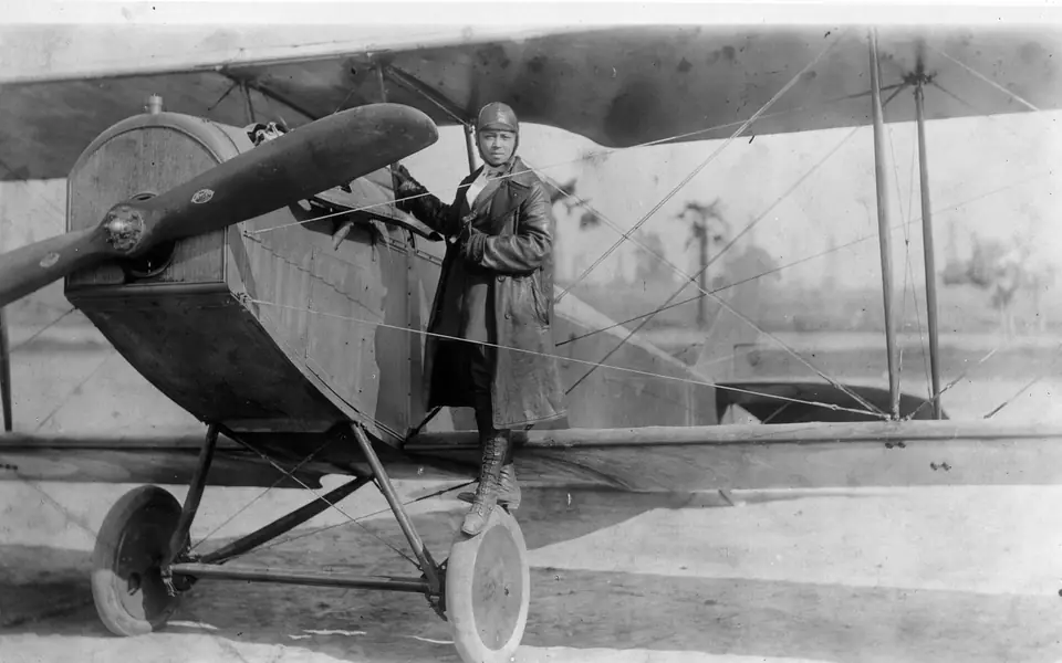 Bessie Coleman in flying clothes standing on the wheel of a Curtiss Jenny biplane in a photograph cataloged as 1922